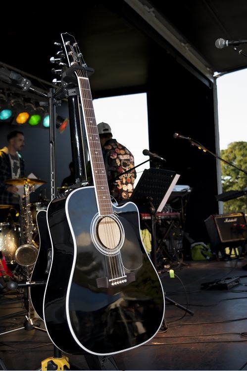 Eine Gitarre, die im Vordergrund auf einer Bühne steht. Im Hintergrund sind Musiker.