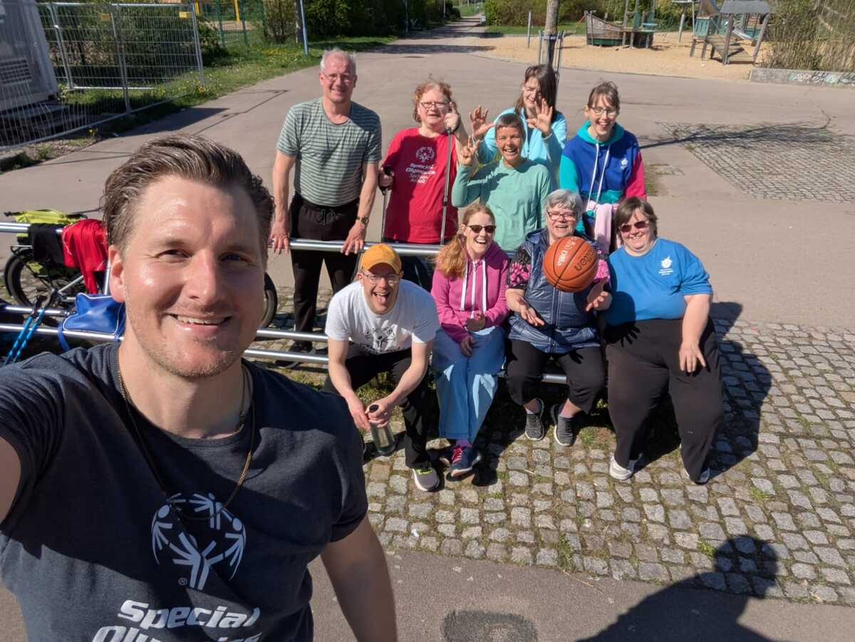 Das Bild zeigt eine Gruppe von motivierten Menschen, die in der Sonne sitzen. Im Vordergrund ist Georg Melzer von Special olympics zu sehen, der das Bild von der Gruppe als Selfie macht
