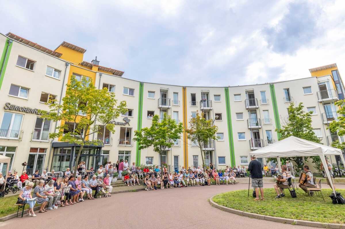 Bewohnerinnen und Bewohner der ProCurand Seniorenresidenz Am Hufeisensee verfolgen im Innenhof ein Konzert mit zwei Akkordeonspielern unter freiem Himmel.