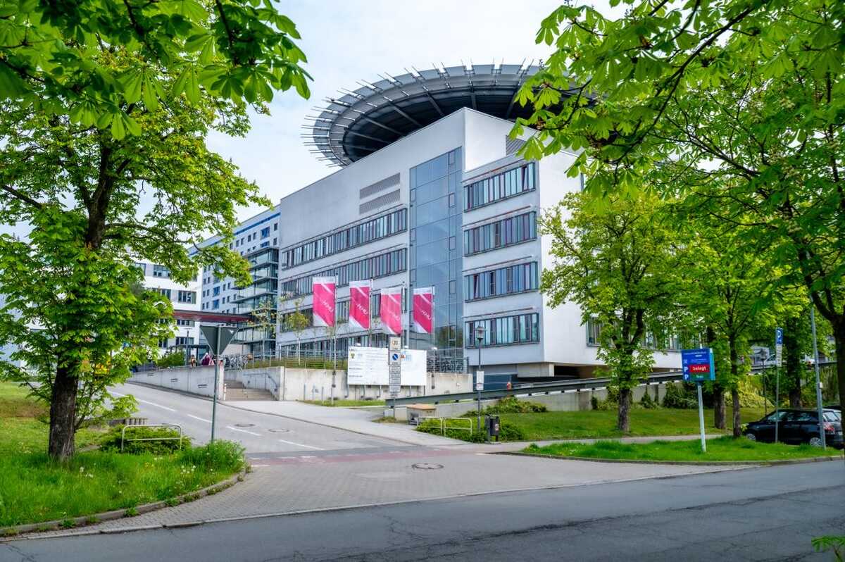 Außenansicht des Universitätsklinikums Halle (Saale). Das moderne Klinikgebäude ist von Bäumen umgeben und dient als Veranstaltungsort der angekündigten Veranstaltung.