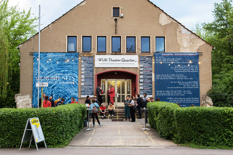 Haupteingang WUK-Theater mit Menschen, die an Stehtischen reden und auf den Eingangsstufen sitzen. Links ein großes Transparent mit der Aufschrift Gesund sein. Rechts und links neben der Tür sind Tafeln mit dem Programm zu sehen. Es ist nichts lesbar.