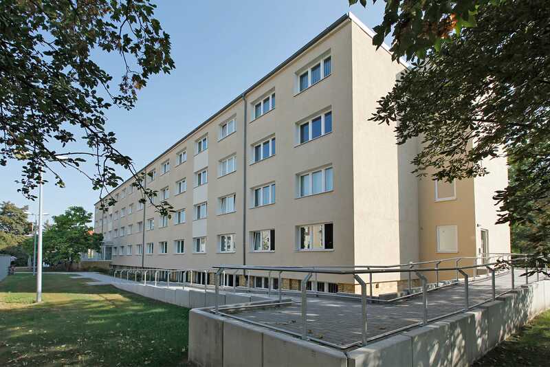 Block mit 4 Etagen, gelber Fassade Flachdach und einer langen Rollstuhlrampe, fast an der ganzen Fassade entlang.