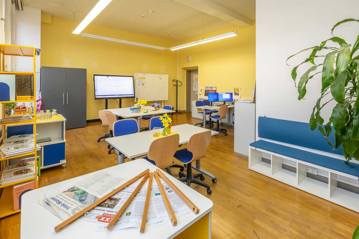 Kleiner freundlich wirkender Klassenraum mit Tafel, Monitor und auf den Tischen verschiedene Unterrichtsmaterialien und 